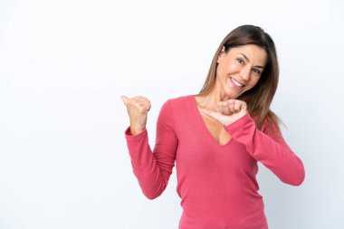 Beyaz arka planda izole edilmiş orta yaşlı beyaz kadın bir ürünü sunmak için kenarı işaret ediyor.