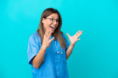 Orta yaşlı cerrah kadın, mavi arka planda, şaşırmış yüz ifadesi ile izole edilmiş.