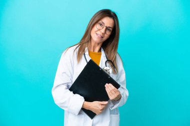 Mavi arka planda izole edilmiş orta yaşlı bir kadın doktor önlüğü giyiyor ve elinde bir klasör tutuyor.