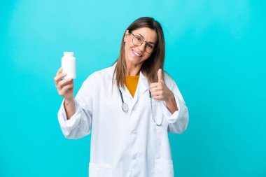 Mavi arka planda izole edilmiş orta yaşlı bir kadın doktor önlüğü giyiyor ve elinde haplar var.