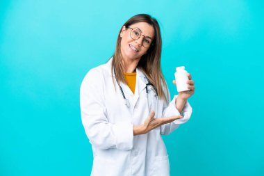 Mavi arka planda izole edilmiş orta yaşlı bir kadın doktor önlüğü giyiyor ve elinde haplar var.