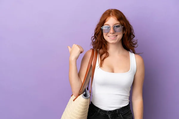 十代の赤毛の女の子は 製品を提示する側を指して紫色の背景に隔離されたビーチバッグを保持 — ストック写真