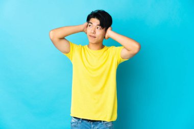 Mavi arka planda izole edilmiş genç bir Çinli hayal kırıklığına uğramış ve kulaklarını kapatmış.