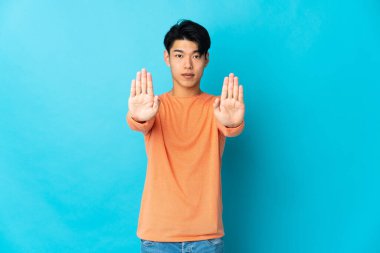 Genç Çinli adam mavi arka planda izole edilmiş. Durma hareketi yapıyor ve hayal kırıklığına uğruyor.