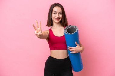 Genç sporcu Ukraynalı kadın yoga derslerine giderken pembe arka planda izole edilmiş bir minderi tutuyor ve parmaklarıyla üçe kadar sayıyor.