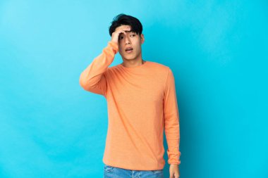 Mavi arka planda izole edilmiş genç bir Çinli yan tarafa bakarken sürpriz jest yapıyor.