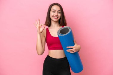 Genç sporcu Ukraynalı kadın yoga derslerine giderken elinde pembe arka planda izole edilmiş bir minder tutarken parmaklarıyla tamam işareti gösteriyor.