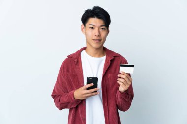 Genç Çinli bir adam beyaz arka planda izole edilmiş cep telefonuyla kredi kartıyla alışveriş yapıyor.