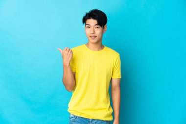 Mavi arka planda izole edilmiş genç Çinli bir adam bir ürün sunmak için kenarı işaret ediyor.