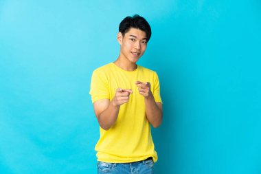 Mavi arka planda izole edilmiş genç bir Çinli şaşırmış ve ön tarafı işaret ediyor.