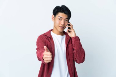 Genç Çinli adam beyaz arka planda izole edildi. Başparmaklarını kaldırırken cep telefonuyla sohbet ediyor.