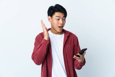 Genç Çinli adam beyaz arka planda izole edilmiş kameraya bakarken şaşırmış bir ifadeyle cep telefonunu kullanıyor.