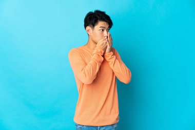 Genç Çinli adam mavi arka planda izole edilmiş ağzını kapatıyor ve yan tarafa bakıyor.