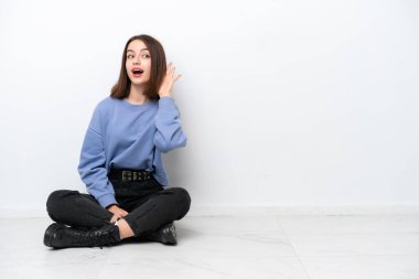 Genç Ukraynalı bir kadın, beyaz arka planda izole bir şekilde yerde oturuyor ve elini kulağına koyarak bir şeyler dinliyor.