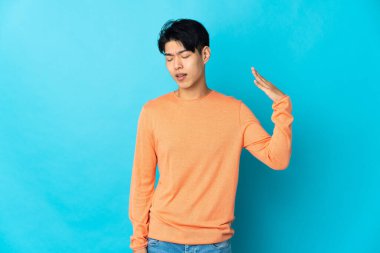 Genç Çinli adam mavi arka planda izole edilmiş, yorgun ve hasta bir ifadeyle.