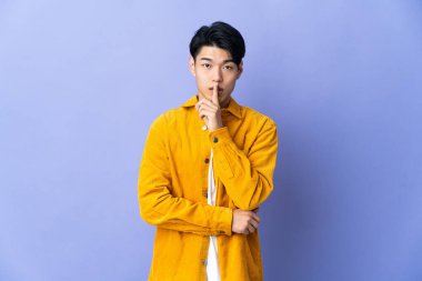 Mor arka planda izole edilmiş genç Çinli bir adam sessizlik işareti gösteriyor. Parmağını ağzına sokuyor.