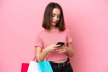 Young Ukrainian woman isolated on pink background holding shopping bags and writing a message with her cell phone to a friend