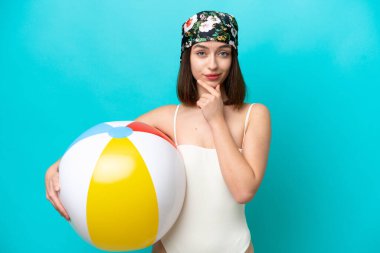 Young Ukrainian woman holding beach ball isolated on blue background thinking