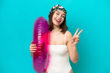 Mavi arka planda hava yatağı tutan genç Ukraynalı kadın gülümsüyor ve zafer işareti gösteriyor.