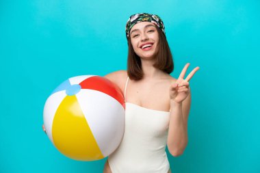 Genç Ukraynalı elinde plaj topu tutan kadın mavi arka planda izole edilmiş gülümsüyor ve zafer işareti gösteriyor.