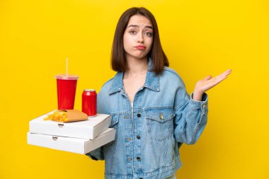 Young Ukrainian woman holding fast food isolated on yellow background having doubts while raising hands