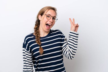 Beyaz arka planda izole edilmiş genç beyaz kadın gülümsüyor ve zafer işareti gösteriyor.