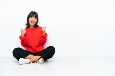 Genç melez kadın, beyaz arka planda soyutlanmış bir şekilde yerde oturuyor. Baş parmakları havada ve gülümsüyor.