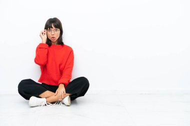 Genç melez kadın, beyaz arka planda gözlüklü ve şaşırmış bir şekilde yerde oturuyor.