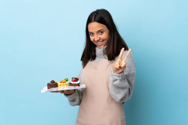 Pasta şefi mavi arka planda büyük bir pasta tutuyor gülümsüyor ve zafer işareti gösteriyor..