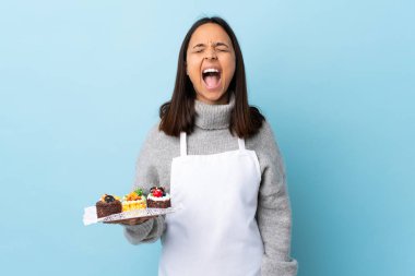 Pasta şefi büyük bir pasta tutuyor mavi arka planda ve ağzı açık bir şekilde öne doğru bağırıyor..
