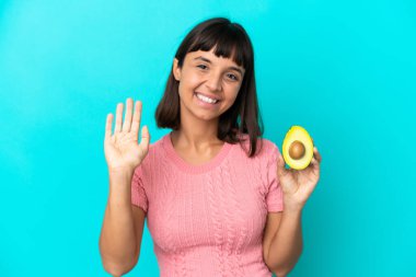 Mavi arka planda avakado tutan genç melez kadın mutlu bir ifadeyle selamlıyor.