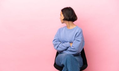 Genç melez kadın arka planda pembe arka planda izole edilmiş bir sandalyede oturuyor.