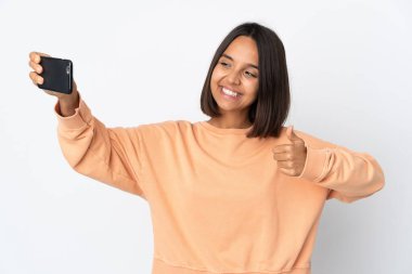 Beyaz arka planda izole edilmiş genç Latin bir kadın cep telefonuyla selfie çekiyor.