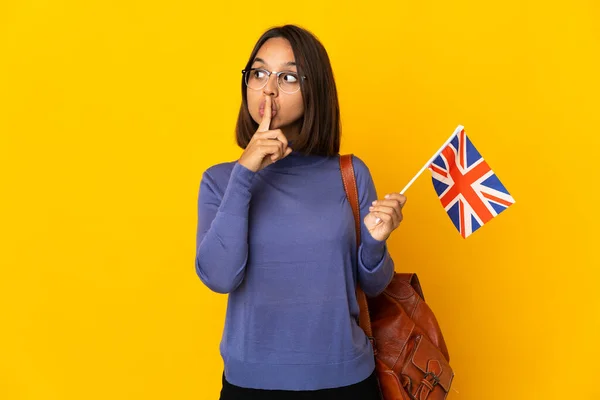 黄色の背景に英国の国旗を掲げた若いラテン女性が口の中に指を入れて沈黙のジェスチャーの兆候を示しています — ストック写真