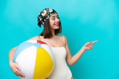 Young Ukrainian woman holding beach ball isolated on blue background pointing finger to the side and presenting a product