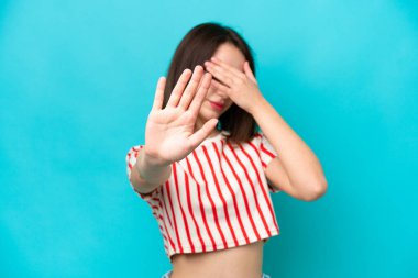 Mavi arka planda izole edilmiş genç Ukraynalı kadın jesti durdurup yüzünü kapatıyor.