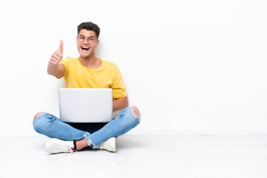 Young man sitting on the floor isolated on white background with thumbs up because something good has happened