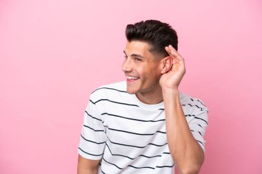 Pembe arka planda izole edilmiş genç beyaz adam elini kulağına koyarak bir şeyler dinliyor.