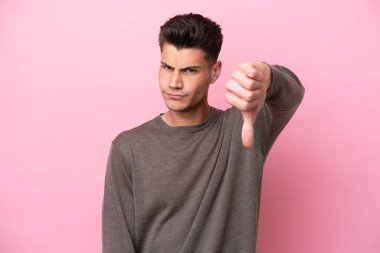 Pembe arka planda izole edilmiş genç beyaz adam olumsuz ifadeyle baş parmağını aşağı gösteriyor.
