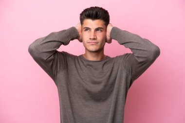 Pembe arka planda izole edilmiş genç beyaz adam hayal kırıklığına uğramış ve kulaklarını kapatmış.