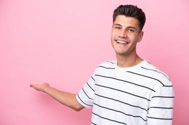 Pembe arka planda izole edilmiş genç beyaz adam, davet edilmek için ellerini yana uzatıyor.