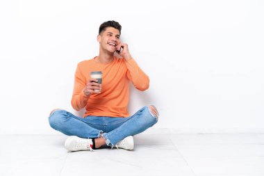Genç adam beyaz arka planda izole bir şekilde yerde oturuyor. Elinde kahve ve cep telefonu var.