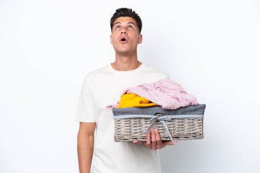 Beyaz arka planda çamaşır sepetini tutan genç beyaz bir adam yukarı bakıyor ve şaşırmış bir ifadeyle