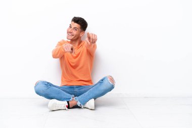 Genç bir adam beyaz arka planda izole bir şekilde yerde oturuyor. Gülümserken seni işaret ediyor.