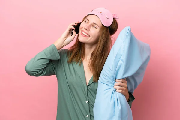 Young English woman in pajamas isolated on pink background in pajamas and holding a pillow while talking to mobile
