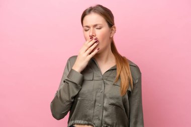 Pembe arka planda izole edilmiş genç beyaz bir kadın esniyor ve geniş bir ağzı eliyle kapatıyor.