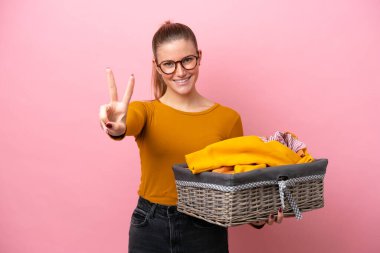 Pembe arka planda izole edilmiş çamaşır sepeti tutan beyaz bir kadın gülümsüyor ve zafer işareti gösteriyor.