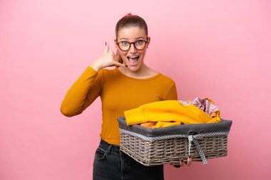 Pembe arka planda izole edilmiş çamaşır sepetini tutan beyaz bir kadın telefon jesti yapıyor. Bana geri dönüş işareti ver.