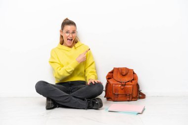 Beyaz arka planda izole bir katta oturan genç beyaz kadın şaşırmış ve işaret ediyor.