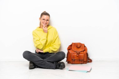 Beyaz arka planda beyaz bir zeminde oturan beyaz tenli genç bir kadın gözlüklü ve gülümsüyor.